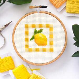 May include: A cross-stitch embroidery hoop featuring a yellow lemon design within a yellow and white gingham border. The embroidery is set in a wooden hoop, with spools of yellow and orange thread and small scissors nearby.