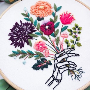 Puede incluir: Arte de bordado en aro que presenta una mano de esqueleto sosteniendo un ramo de flores coloridas. Las flores son rosas, rojas, moradas, naranjas y verdes. El fondo es blanco.