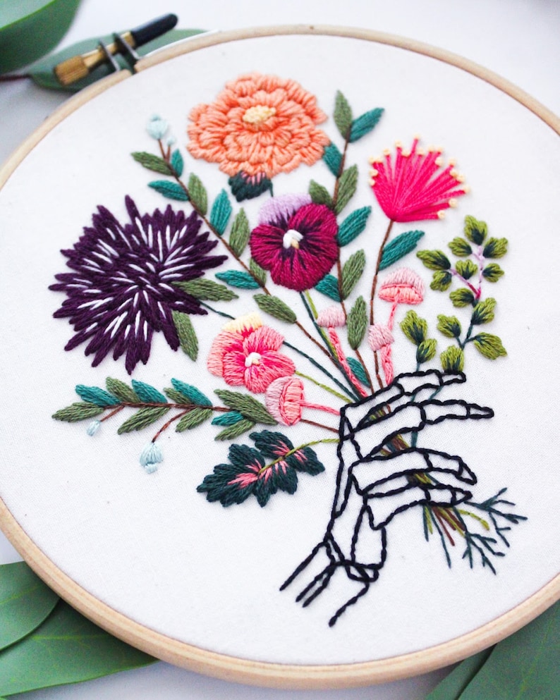 Puede incluir: Arte de bordado en aro que presenta una mano de esqueleto sosteniendo un ramo de flores coloridas. Las flores son rosas, rojas, moradas, naranjas y verdes. El fondo es blanco.