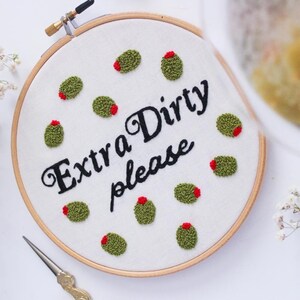 May include: Embroidered hoop art with the text "Extra Dirty please" and green olives with red centers.