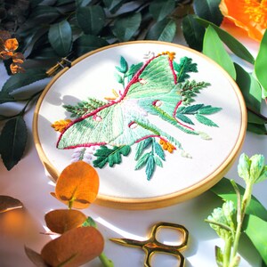 May include: Embroidered hoop art featuring a green luna moth with pink and orange accents surrounded by green foliage.