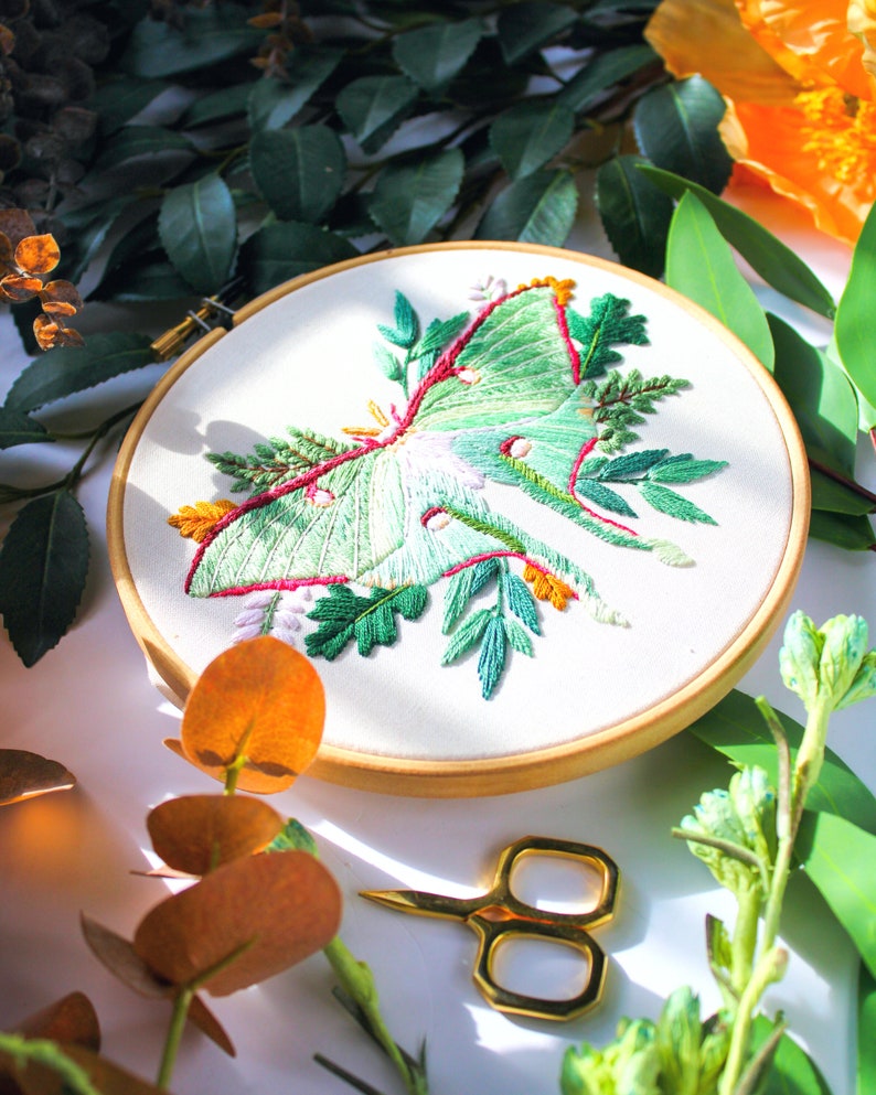 May include: Embroidered hoop art featuring a green luna moth with pink and orange accents surrounded by green foliage.