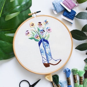 May include: A wooden embroidery hoop with a white fabric background. The fabric has an embroidered design of a blue cowboy boot with a bouquet of colorful pansies inside. The hoop is surrounded by embroidery floss in various colors.