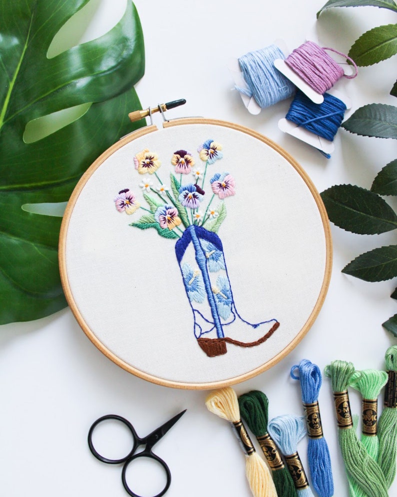 May include: A wooden embroidery hoop with a white fabric background. The fabric has an embroidered design of a blue cowboy boot with a bouquet of colorful pansies inside. The hoop is surrounded by embroidery floss in various colors.
