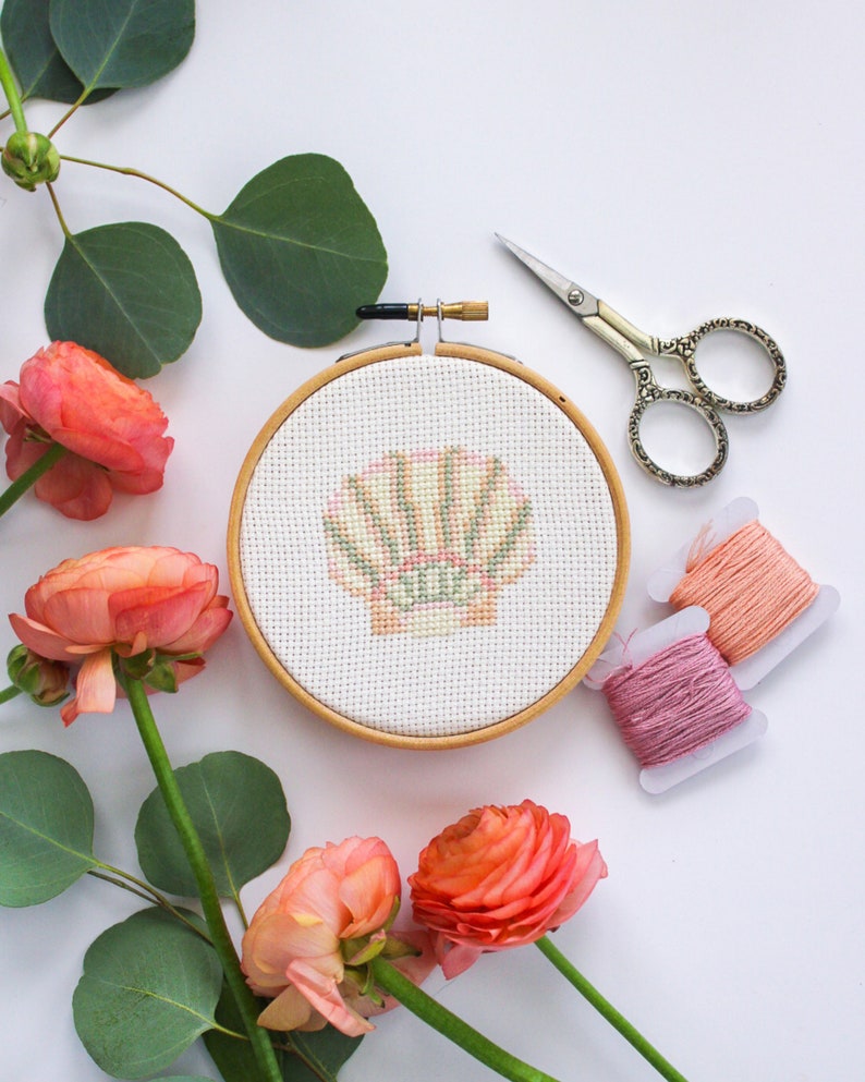 K&ouml;nnte beinhalten: Nahaufnahme eines fertigen Stickrahmens mit einem rosa-gr&uuml;nen Muschelmotiv. Der Rahmen ist von rosa Bl&uuml;ten und gr&uuml;nen Bl&auml;ttern umgeben. Zwei Spulen Stickgarn sind ebenfalls zu sehen.