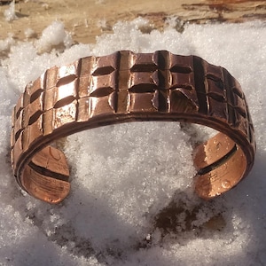 May include: A copper cuff bracelet with a textured, geometric pattern. The open cuff design features a series of raised squares and rectangles, creating a grid-like appearance. The bracelet is displayed on a snowy surface, highlighting its metallic sheen.