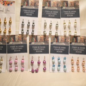 May include: A collection of handmade earrings displayed on cards. The earrings feature various bead designs in gold, silver, pearl, pink, purple, and blue. The cards read "THIS IS DEBS CRAFTING ROOM" and "Thank You".