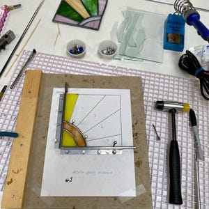 Op de afbeelding: Een lopend glas-in-loodproject, met een stuk glas dat wordt gesneden en een hamer en andere gereedschappen die op een wit rooster oppervlak zijn gelegd. Het glas wordt gesneden op een vel papier met een geel en oranje ontwerp, met de tekst "#1 easiest option all around".
