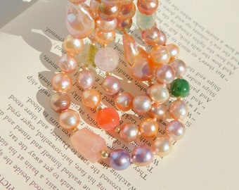 May include: A multi-strand pearl necklace with pink, peach, and white pearls. The necklace also features small, colorful gemstones and a few larger, irregular-shaped pink stones.