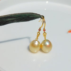 以下が含まれることがあります： 金色のイヤリングのペア。イヤリングには、それぞれ金色の真珠のドロップが1つずつ付いています。真珠は明るい金色で、イヤリングにはチェーンに小さな赤い宝石が付いています。