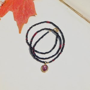 Könnte beinhalten: Eine mehrreihige Halskette mit einem goldenen Anhänger mit einem rosa Edelstein. Die Halskette besteht aus kleinen, dunkelblauen und roten Perlen. Der Anhänger ist oval und in einen goldenen Rahmen gefasst. Die Halskette wird auf einer weißen Oberfläche präsentiert.