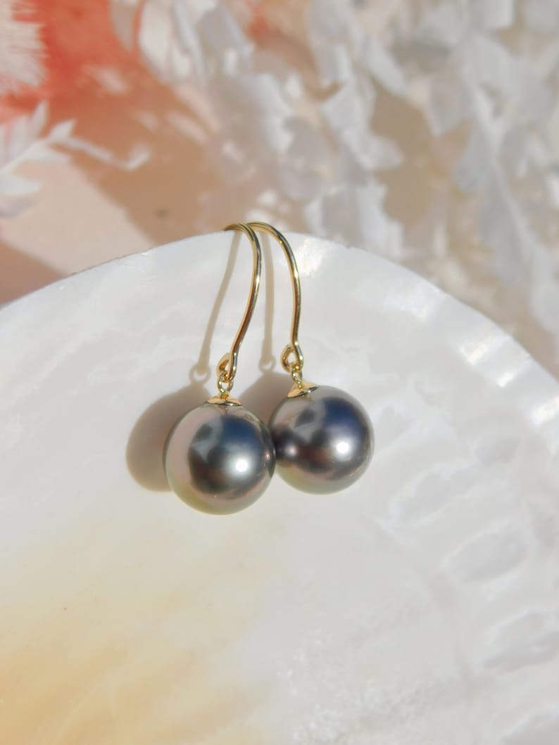 May include: A pair of black pearl drop earrings with gold hooks. The pearls are round and have a slight iridescent sheen. The earrings are displayed on a white seashell.
