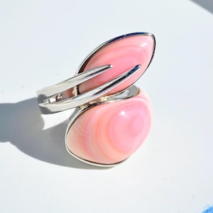 Könnte beinhalten: Ein offener Ring mit zwei glatten, polierten Cabochons aus rosafarbener Muschelschale, die in eine silberfarbene Metallschiene gefasst sind. Die Steine zeigen natürliche konzentrische Bänderungen in verschiedenen hellen Rosatönen.