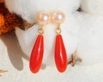 May include: A pair of drop earrings featuring round, cream-colored pearls attached to gold-toned metal jump rings, which connect to smooth, elongated, teardrop-shaped bright red coral pendants that hang vertically.