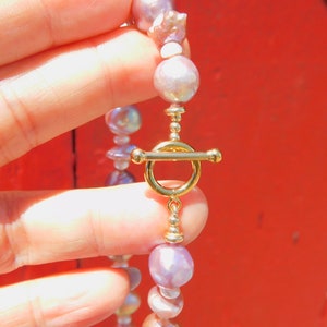 May include: A delicate necklace made of iridescent purple and white pearls with a gold clasp. The necklace is held in a hand against a red background.