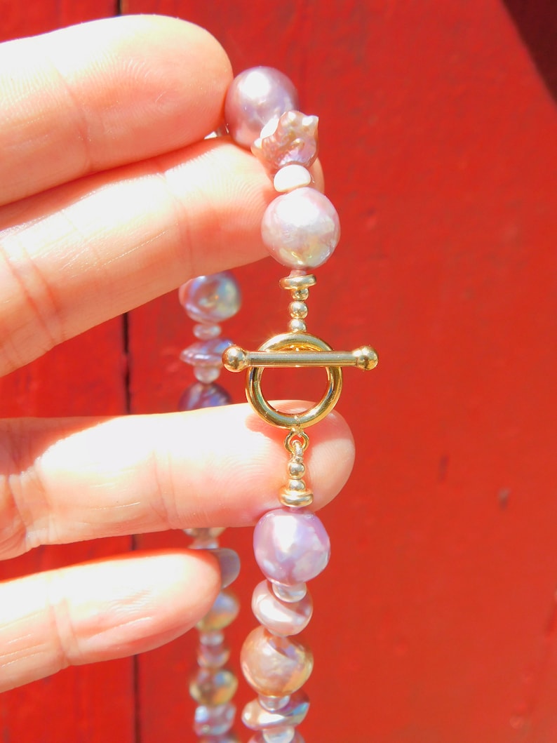 May include: A delicate necklace made of iridescent purple and white pearls with a gold clasp. The necklace is held in a hand against a red background.
