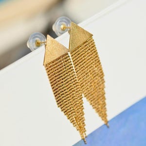 May include: A pair of gold-toned dangle earrings featuring a triangular top piece with a brushed texture. Attached to the base of each triangle is a long, cascading fringe made of multiple strands of tiny, reflective gold-coloured metal beads.