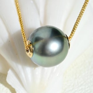 May include: A round, dark gray Tahitian pearl pendant suspended on a thin, gold-colored chain. The pearl features a smooth, lustrous surface with subtle green and blue undertones, held in place by a small, polished gold-toned metal bail.