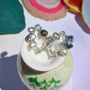 May include: Silver earrings featuring irregular grey and white baroque pearls. The earrings are presented on a small, white ceramic dish, placed on a green and white decorative vase. Agate slices in the background.
