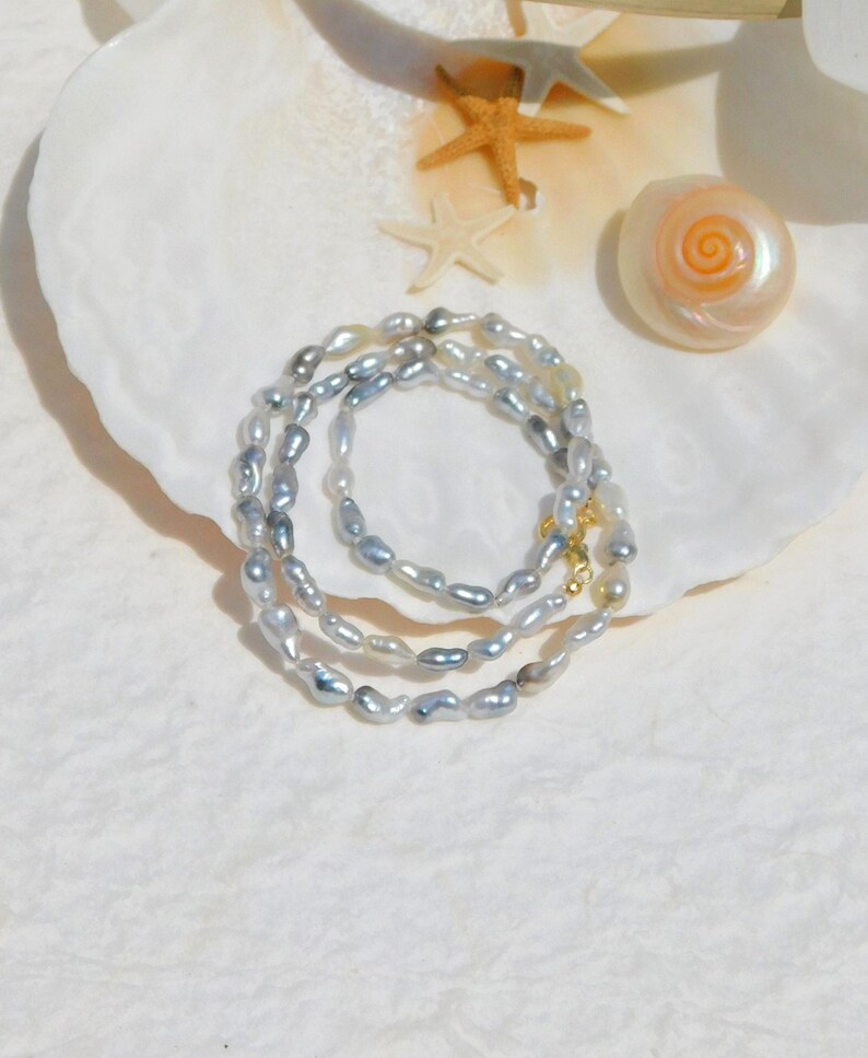 Op de afbeelding: Een ketting bestaande uit meerdere strengen onregelmatige, glanzende grijze parels. De parels zijn geregen in een doorlopende lus, voorzien van een kleine, gepolijste goudkleurige metalen sluiting die de uiteinden met elkaar verbindt.