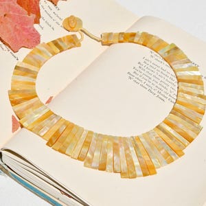 May include: A striking necklace made from rectangular, iridescent, yellow-orange shell segments. The necklace is laid open on a book with text. The clasp is a round, shell-like button. A distinctive piece of jewellery.