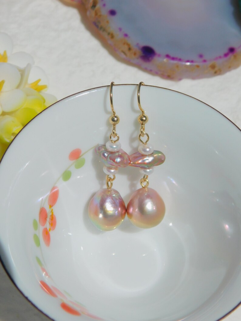 May include: A pair of gold dangle earrings with pink and white baroque pearls. The pearls are iridescent and have a pearlescent sheen.