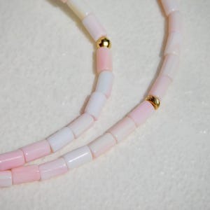 May include: A delicate necklace featuring alternating pink and white beads. The beads are strung together with gold-colored accents. The necklace is displayed on a white, textured surface, highlighting the soft colors and elegant design.