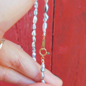 Op de afbeelding: Een ketting met onregelmatige, langwerpige witte en grijze parels. De ketting is voorzien van een goudkleurige ronde veerringsluiting met een kleine blauwe accentsteen. Het metalen onderdeel is duidelijk gestempeld met de tekst Au750.