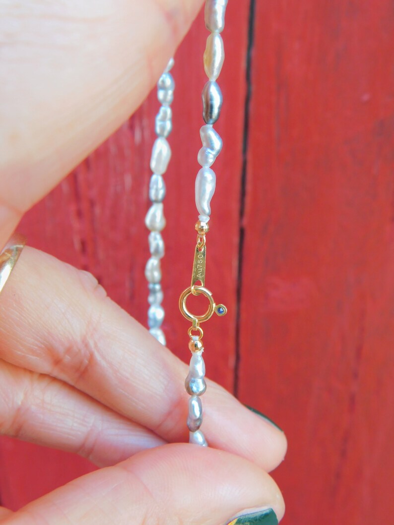 Op de afbeelding: Een ketting met onregelmatige, langwerpige witte en grijze parels. De ketting is voorzien van een goudkleurige ronde veerringsluiting met een kleine blauwe accentsteen. Het metalen onderdeel is duidelijk gestempeld met de tekst Au750.