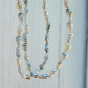 Op de afbeelding: Een lange ketting met meerdere strengen onregelmatige, barokvormige parels in tinten wit, cr&egrave;me en zachtgrijs. De parels hebben een glanzend, reflecterend oppervlak en zijn aan elkaar geregen voor een trapsgewijze, gelaagde uitstraling.