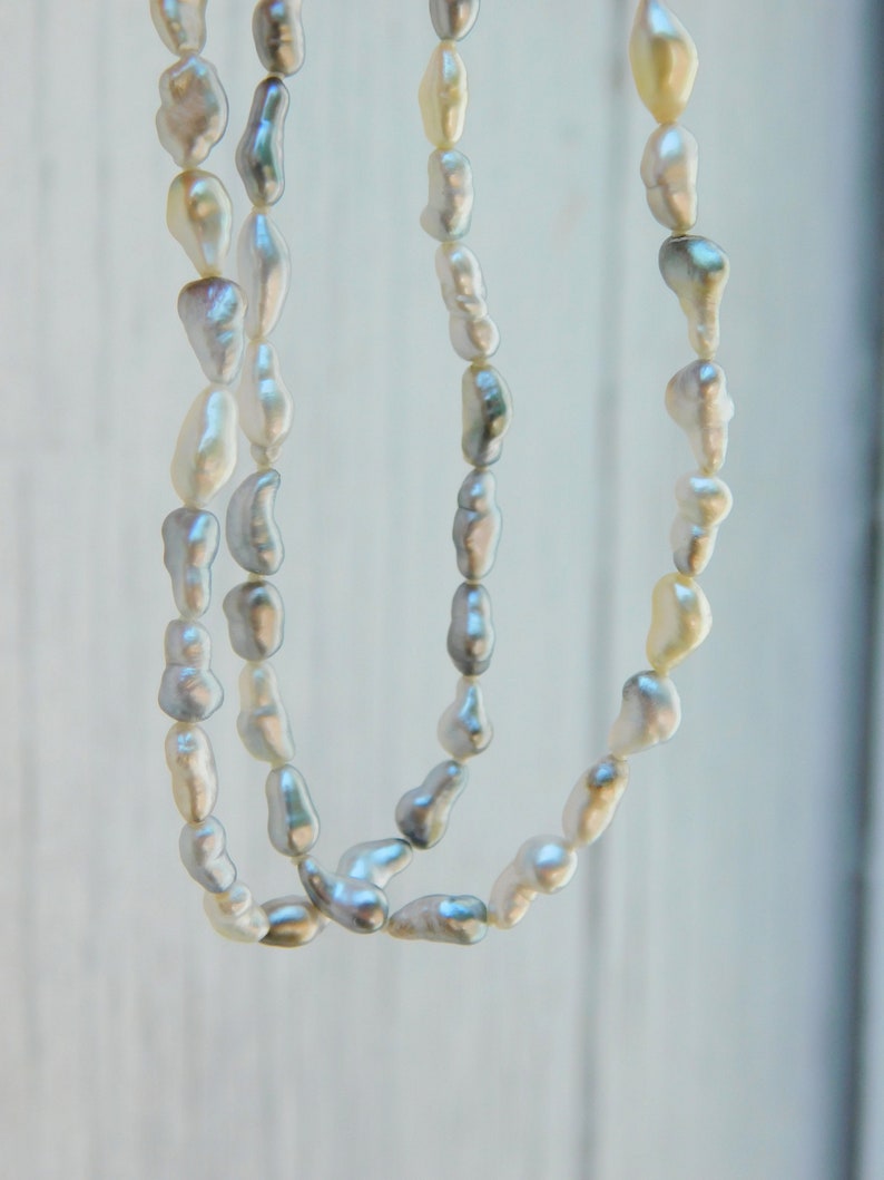 Op de afbeelding: Een lange ketting met meerdere strengen onregelmatige, barokvormige parels in tinten wit, cr&egrave;me en zachtgrijs. De parels hebben een glanzend, reflecterend oppervlak en zijn aan elkaar geregen voor een trapsgewijze, gelaagde uitstraling.