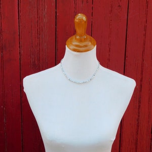 Op de afbeelding: Een delicate ketting met kleine, lichtblauwe kralen gerangschikt in een enkele streng. De ketting wordt getoond op een witte etalagepop-torso, wat de subtiele kleur en uniforme vorm van de individuele kralen over de hele lengte benadrukt.