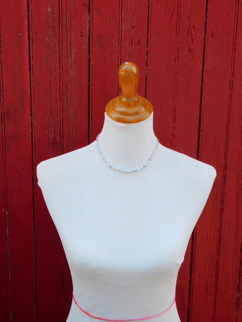Op de afbeelding: Een delicate ketting met kleine, lichtblauwe kralen gerangschikt in een enkele streng. De ketting wordt getoond op een witte etalagepop-torso, wat de subtiele kleur en uniforme vorm van de individuele kralen over de hele lengte benadrukt.