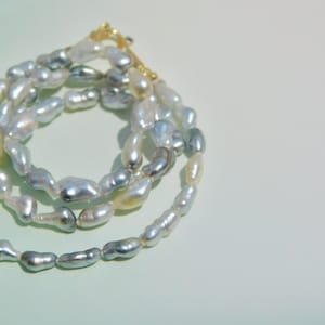 Op de afbeelding: Een ketting bestaande uit onregelmatige, barokke parels in iriserende tinten zilver, grijs en cr&egrave;me. De streng is voorzien van een goudkleurige metalen sluiting aan de bovenzijde, die de uiteinden van de parelketting in een cirkel verbindt.