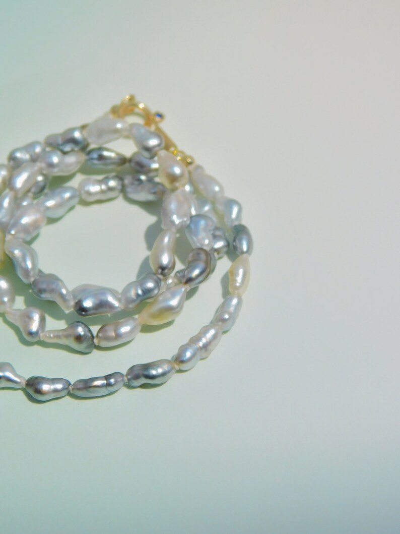 Op de afbeelding: Een ketting bestaande uit onregelmatige, barokke parels in iriserende tinten zilver, grijs en cr&egrave;me. De streng is voorzien van een goudkleurige metalen sluiting aan de bovenzijde, die de uiteinden van de parelketting in een cirkel verbindt.