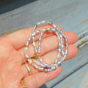 Op de afbeelding: Een ketting bestaande uit kleine, onregelmatige, glanzende witte en grijze parels. De ketting heeft een goudkleurige ronde sluiting met de tekst AU750 gegraveerd op een klein labeltje, samen met een minuscuul blauw accentsteentje.
