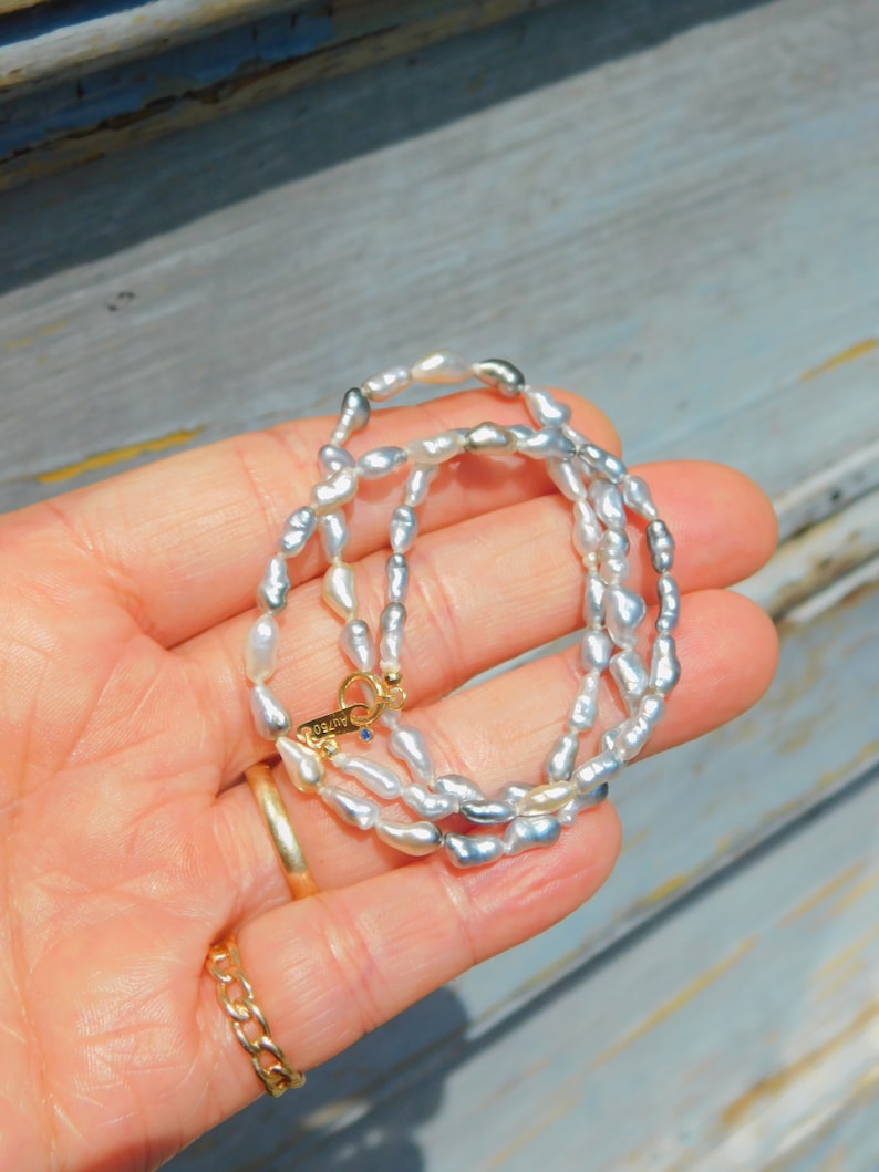 Op de afbeelding: Een ketting bestaande uit kleine, onregelmatige, glanzende witte en grijze parels. De ketting heeft een goudkleurige ronde sluiting met de tekst AU750 gegraveerd op een klein labeltje, samen met een minuscuul blauw accentsteentje.