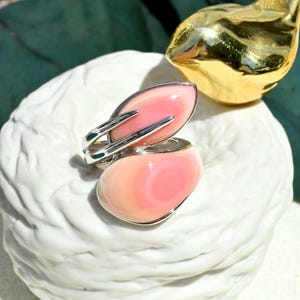 May include: A silver ring featuring two smooth, polished pink conch shell cabochons. The upper stone is an elongated oval shape, while the lower stone is a larger, rounded oval. The silver band wraps around the stones in a bypass style design.