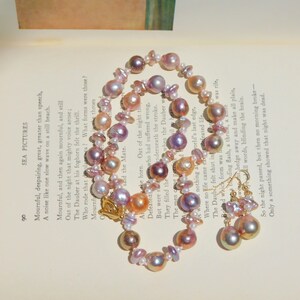 May include: A delicate pearl necklace and earrings set with iridescent pink, purple, and gold tones. The necklace is made of various sizes and shapes of pearls, and the earrings are teardrop shaped.