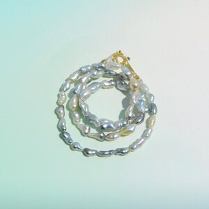 Op de afbeelding: Een ketting bestaande uit onregelmatige, glanzende barokparels in tinten wit, zilver en grijs. Het snoer is in een cirkelvormige opstelling gelegd en is voorzien van een kleine, goudkleurige metalen sluiting met een minuscuul blauw steentje.