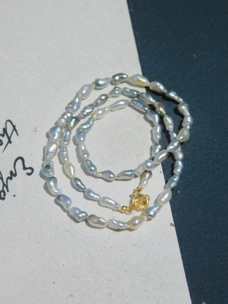 Op de afbeelding: Een ketting bestaande uit talloze kleine, onregelmatige, glanzende grijze en witte barokparels. Het sieraad is voorzien van een ronde goudkleurige metalen knevelsluiting die de twee uiteinden van de parelketting stevig met elkaar verbindt.