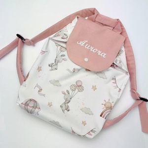 Può includere: Zaino con coulisse rosa e bianco con tasca a patta ricamata con il nome "Aurora". Lo zaino presenta un motivo ripetuto di coniglietti con palloncini, nuvole, stelle e un arcobaleno. Le cinghie e la tasca sono di colore rosa.