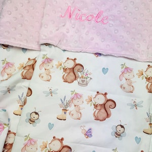 Può includere: Una copertina per neonati rosa e bianca con uno sfondo bianco e un motivo di scoiattoli, coniglietti, fiori e farfalle. La copertina ha un bordo rosa con il nome "Nicole" ricamato.
