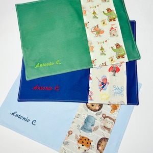 Può includere: Tre tovagliette all'americana con design diversi. La tovaglietta in alto è verde con un bordo bianco e un motivo di volpi, orsi e castelli. La tovaglietta al centro è blu con un bordo bianco e un motivo di supereroi. La tovaglietta in basso è azzurra con un bordo bianco e un motivo di animali. Tutte e tre le tovagliette hanno il nome "Antonio C." ricamato.
