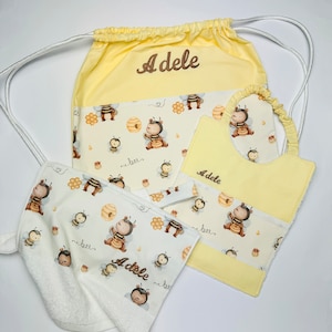 Può includere: Una borsa a cordoncino gialla con una stampa a tema api bianca e gialla e il nome "Adele" ricamato su di essa. La borsa viene fornita con un asciugamano bianco abbinato e un bavaglino giallo con la stessa stampa di api.