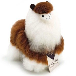 Peut inclure: Une peluche alpaga avec une fourrure épaisse et duveteuse blanche et brune. Le jouet présente une tache brune sur la tête, des oreilles sombres et des yeux noirs. Une étiquette attachée sur le côté affiche le texte INKARI NATURAL ALPACA COMFORT.