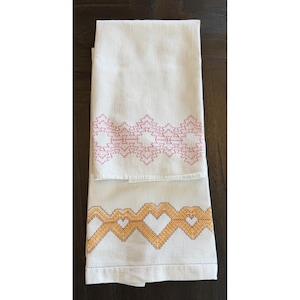 Op de afbeelding: Twee witte linnen handdoeken met geborduurde geometrische patronen. Eén handdoek heeft een roze zigzagontwerp, de andere een oranje hartvormig patroon. De handdoeken zijn opgevouwen en tentoongesteld op een donker houten oppervlak.