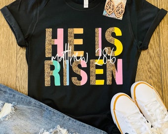 Könnte beinhalten: Schwarzes T-Shirt mit dem Text "HE IS RISEN" in bunten Buchstaben mit einem Leopardenmuster. Der Text "Matthew 28:6" ist in kleineren Buchstaben unter dem Haupttext geschrieben.