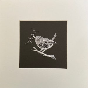 May include: White lace artwork depicting a bird perched on a branch. The bird is detailed with intricate lacework and appears to be holding a twig in its beak.