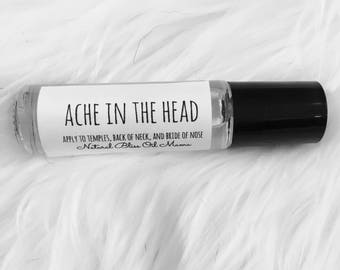 May include: A white roller bottle with a black cap. The label reads "ACHE IN THE HEAD" and "APPLY TO TEMPLES, BACK OF NECK, AND BRIDGE OF NOSE" with the text "Natural Bliss Oil Mama" below.